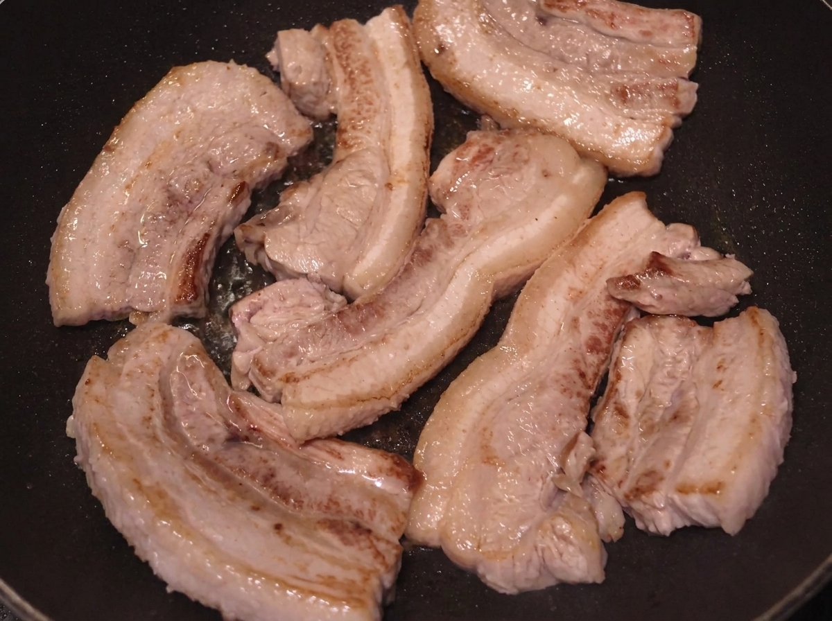 Plusieurs tranches de poitrine de porc en train de dorer à la poêle, avec des surfaces bien colorées.