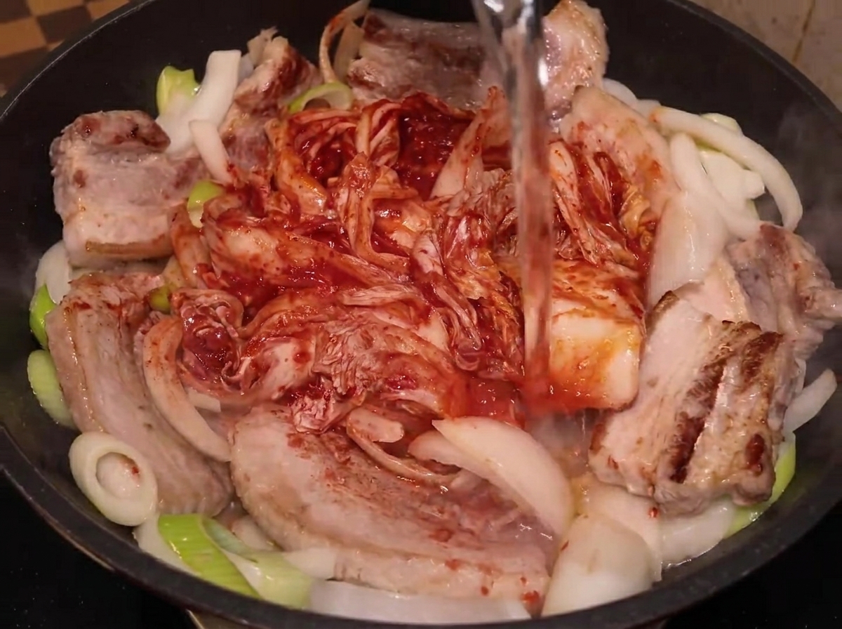 De leau versée depuis une bouteille dans la poêle contenant le porc, les oignons et le kimchi.