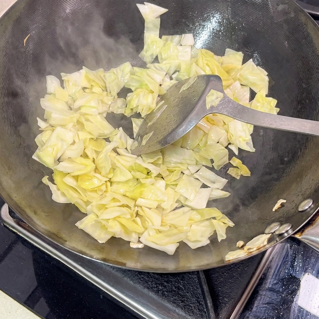 Spatule en métal remuant du chou mariné dans un wok noir très chaud, avec de la vapeur visible.