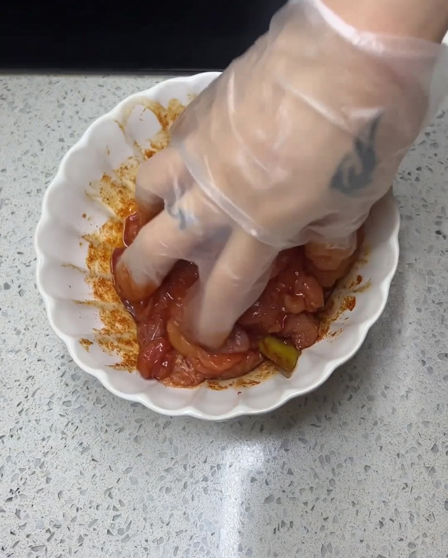 Une main gantée massant une marinade rouge épicée sur des morceaux de poulet.
