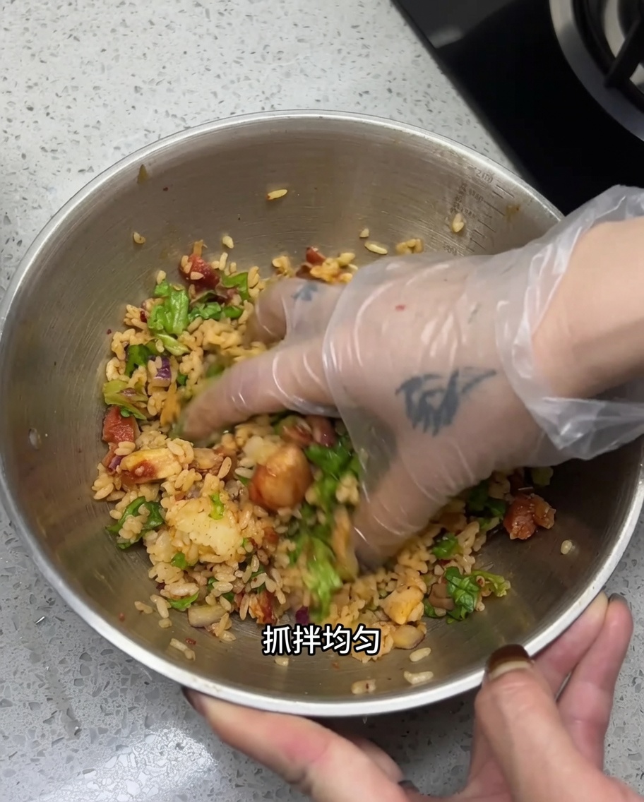 Une main gantée mélangeant le riz, le poulet coupé et les légumes dans un grand bol en acier inoxydable.