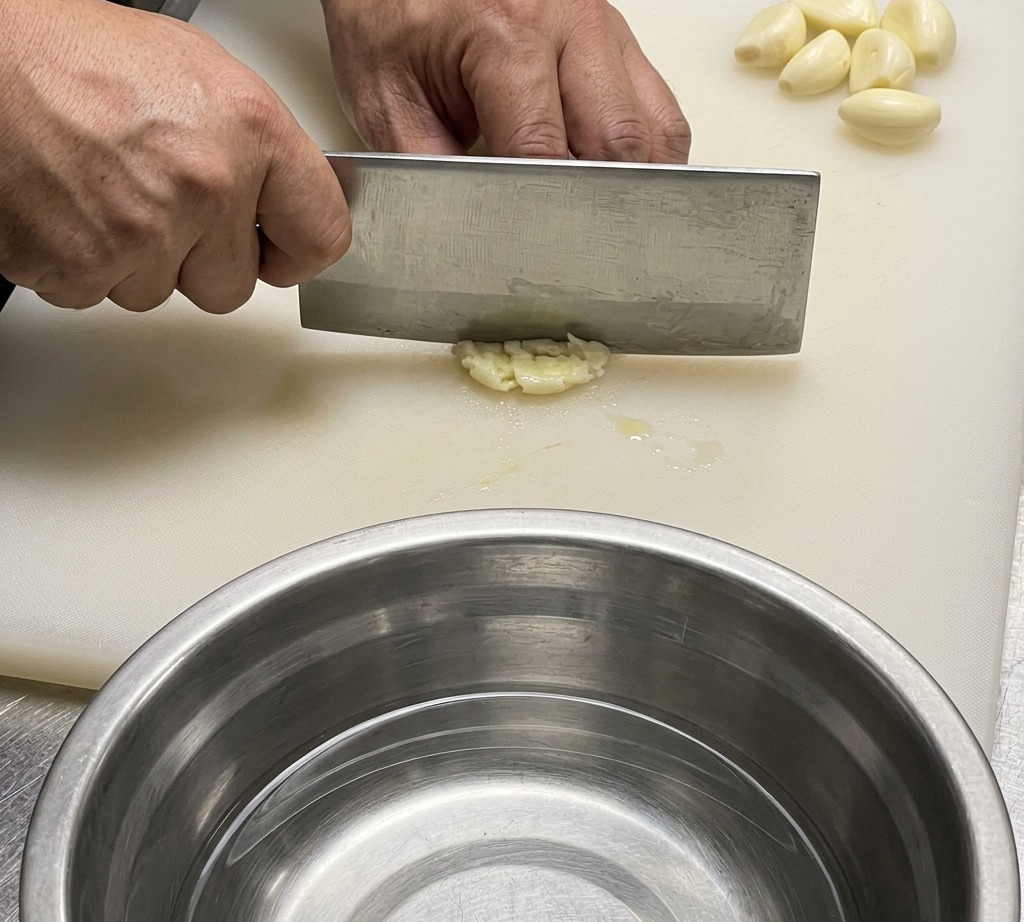 Gros plan dun couteau de cuisine coupant de lail et du gingembre sur une planche à découper blanche.