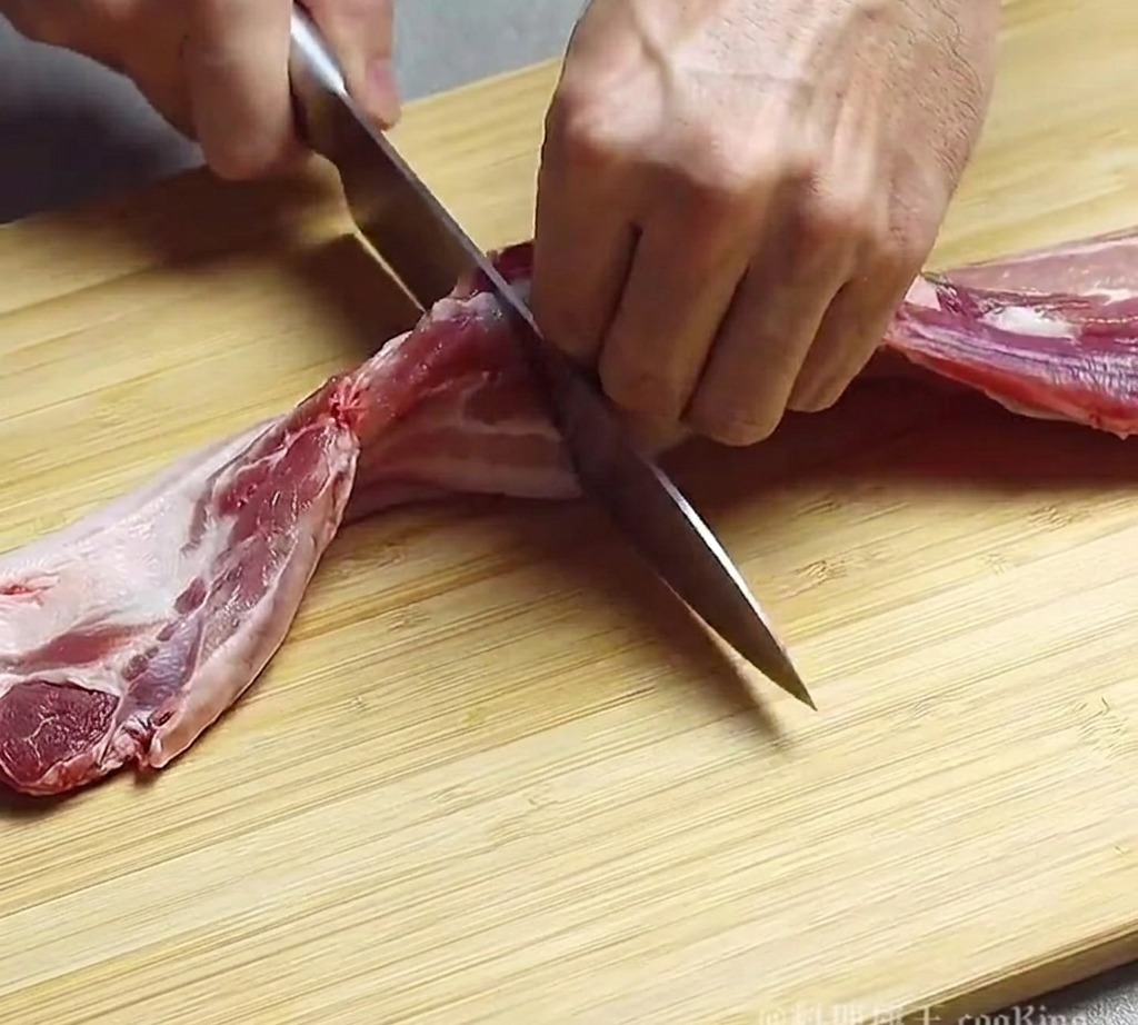Une personne utilisant un couteau de chef pour couper un morceau de poitrine de porc crue en tranches épaisses sur une planche en bois.