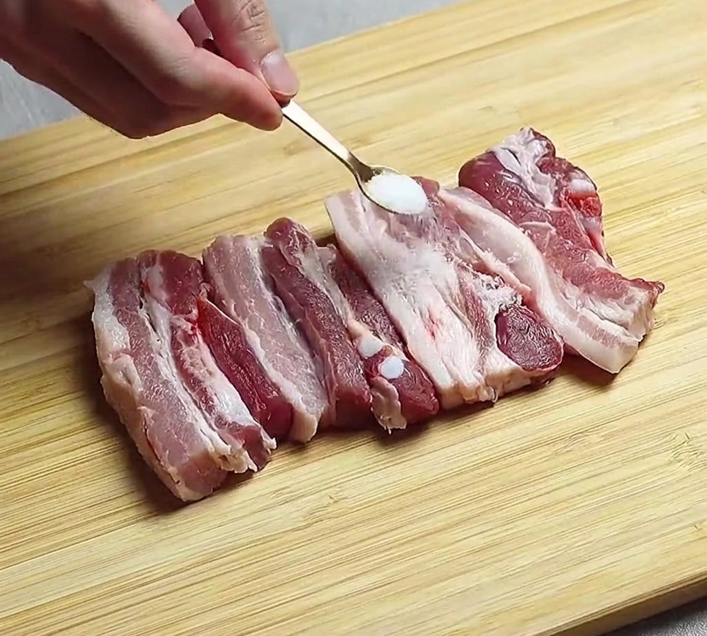 Des tranches de poitrine de porc crue disposées sur une planche en bois en train dêtre assaisonnées avec une pincée de sel.