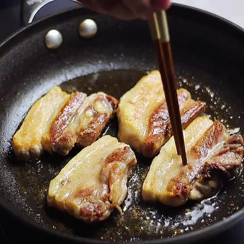 Des tranches de poitrine de porc dorées en train de cuire dans une poêle, avec une baguette utilisée pour vérifier si la viande est cuite.