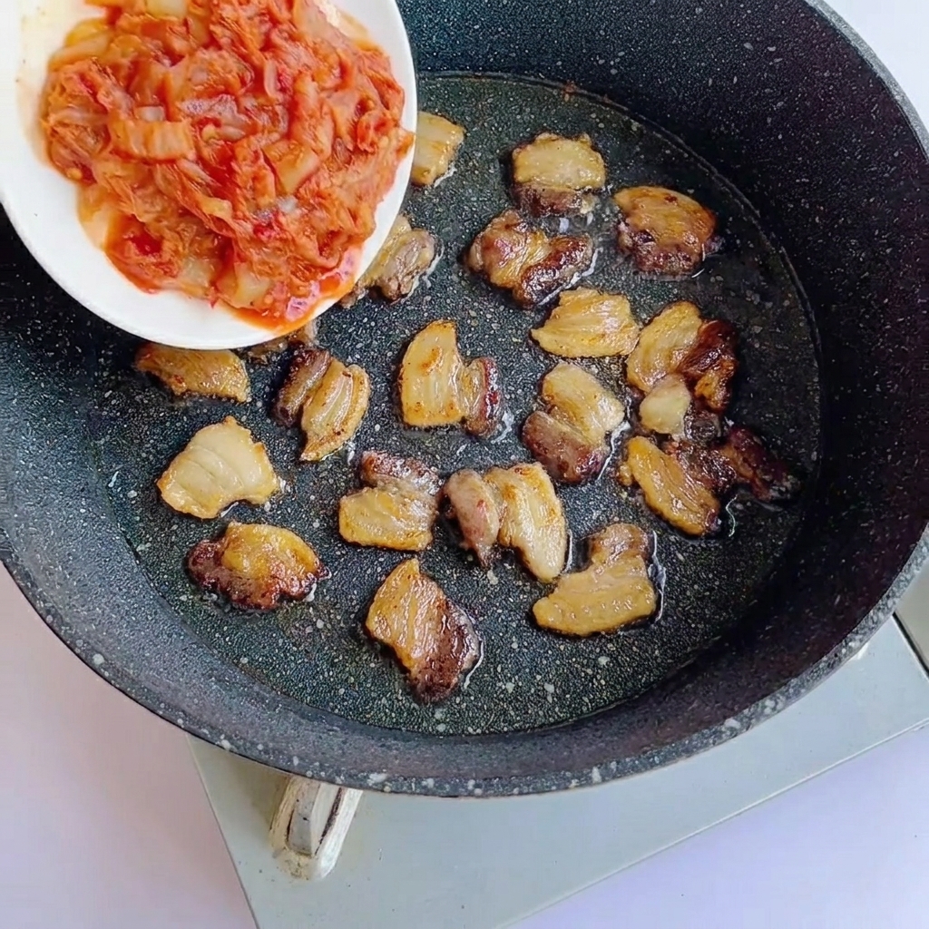 Du kimchi haché ajouté dans une poêle contenant des tranches de porc dorées.