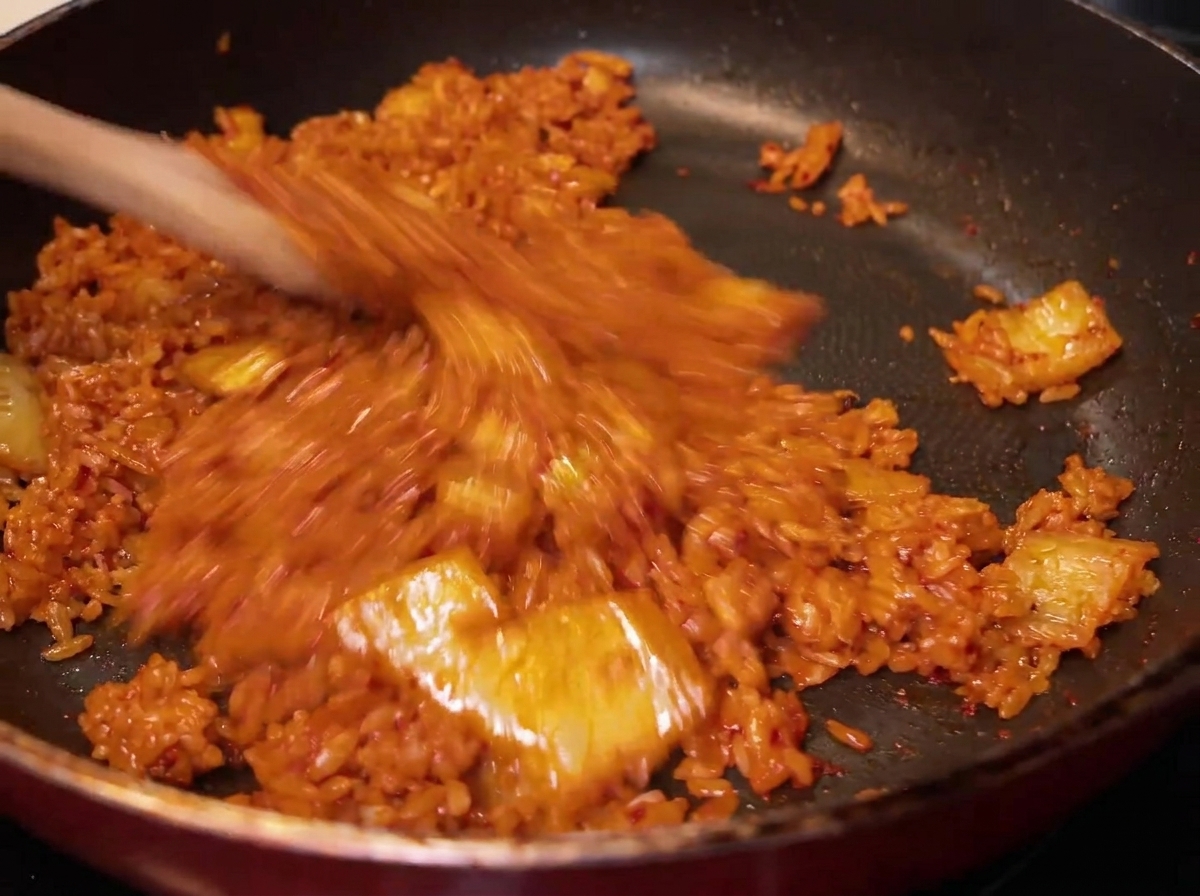 Gros plan sur le riz mélangé au kimchi dans la poêle, prenant une couleur orange uniforme.