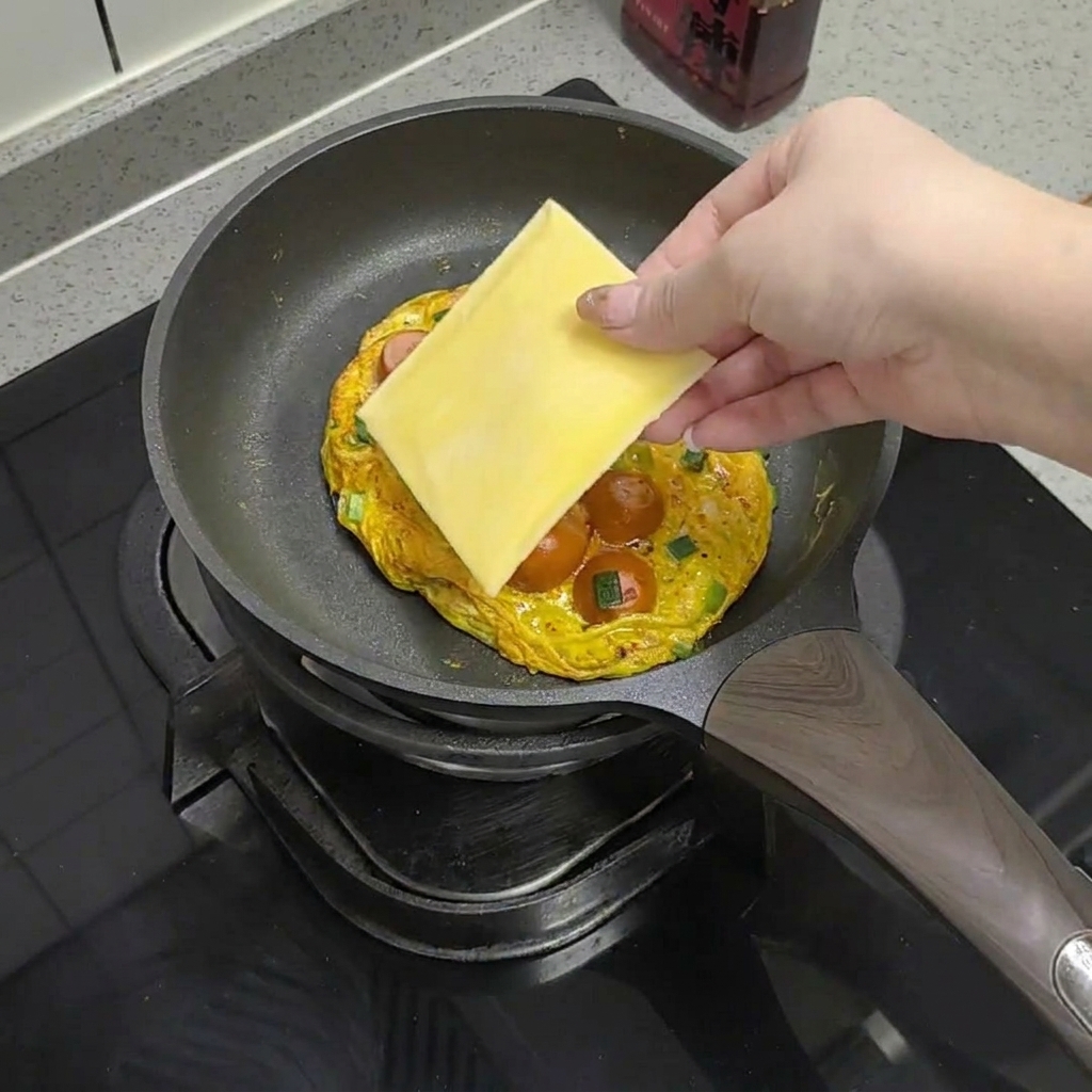 Une tranche de fromage jaune posée sur lomelette aux saucisses dans la poêle.
