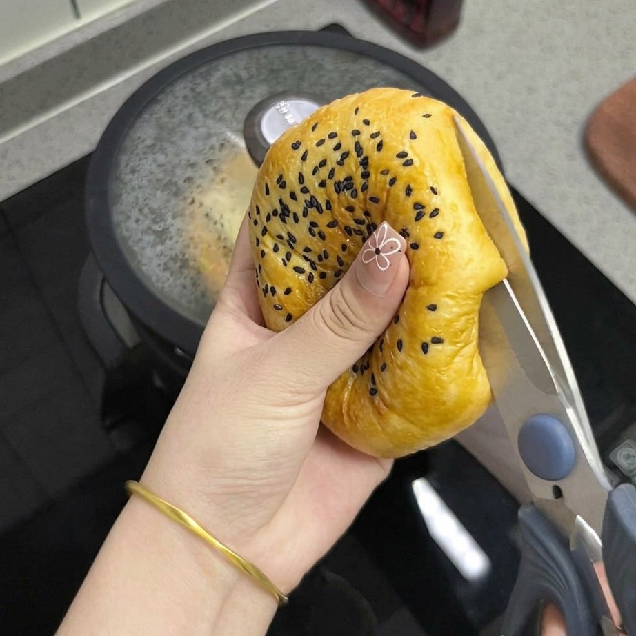 Gros plan sur une main utilisant des ciseaux de cuisine pour couper en deux un pain au sésame doré.