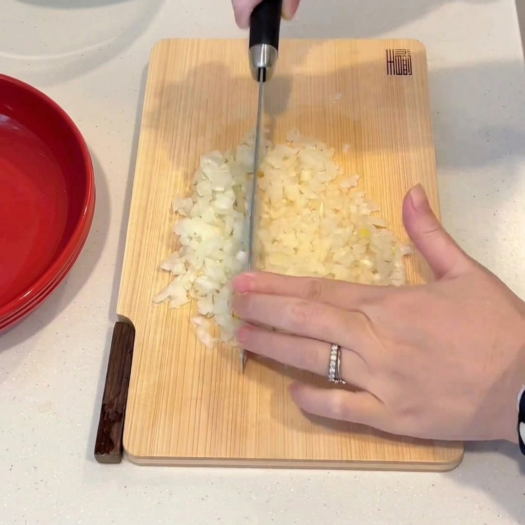 Une main utilisant un couteau de chef pour hacher finement un oignon sur une planche à découper en bois.