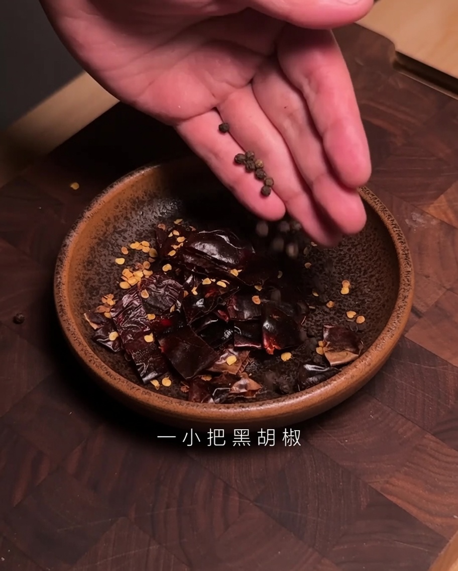 Des mains ajoutant des grains de poivre noir dans un bol contenant des morceaux de piment Guajillo coupé.