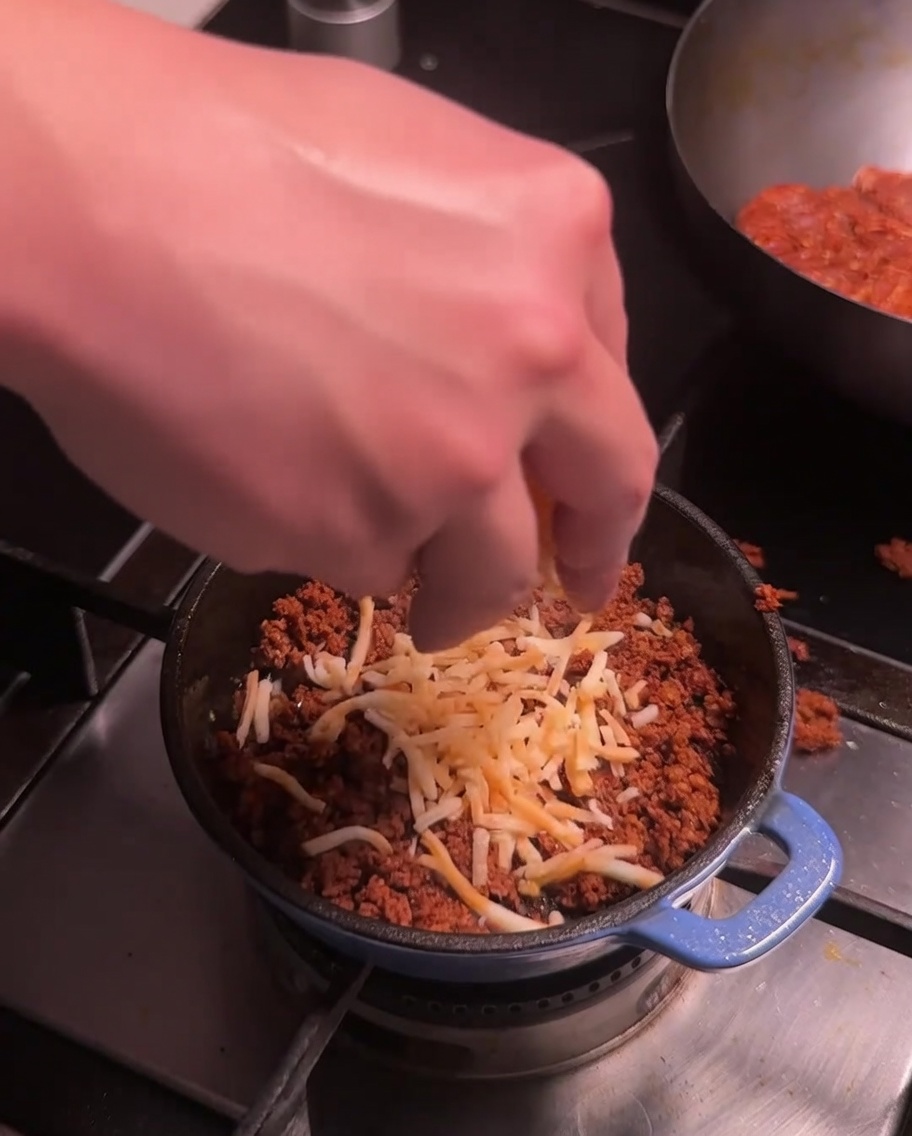 Du fromage râpé étant saupoudré sur la viande de chorizo cuite dans une petite casserole bleue sur la cuisinière.