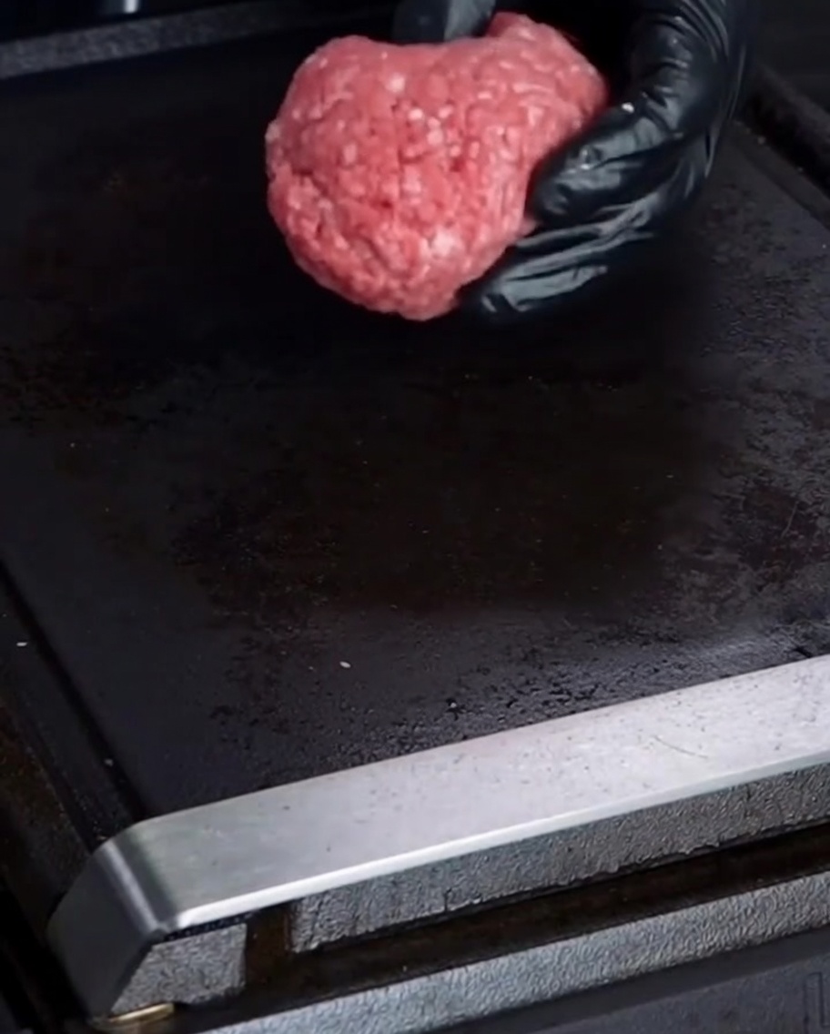 Une main gantée plaçant une boule de bœuf haché cru 75/25 sur une plancha noire.
