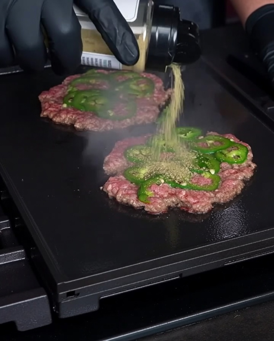 Une main gantée saupoudrant lassaisonnement sur de fines galettes de bœuf écrasées et incrustées de tranches de jalapeño frais sur un gril chaud.