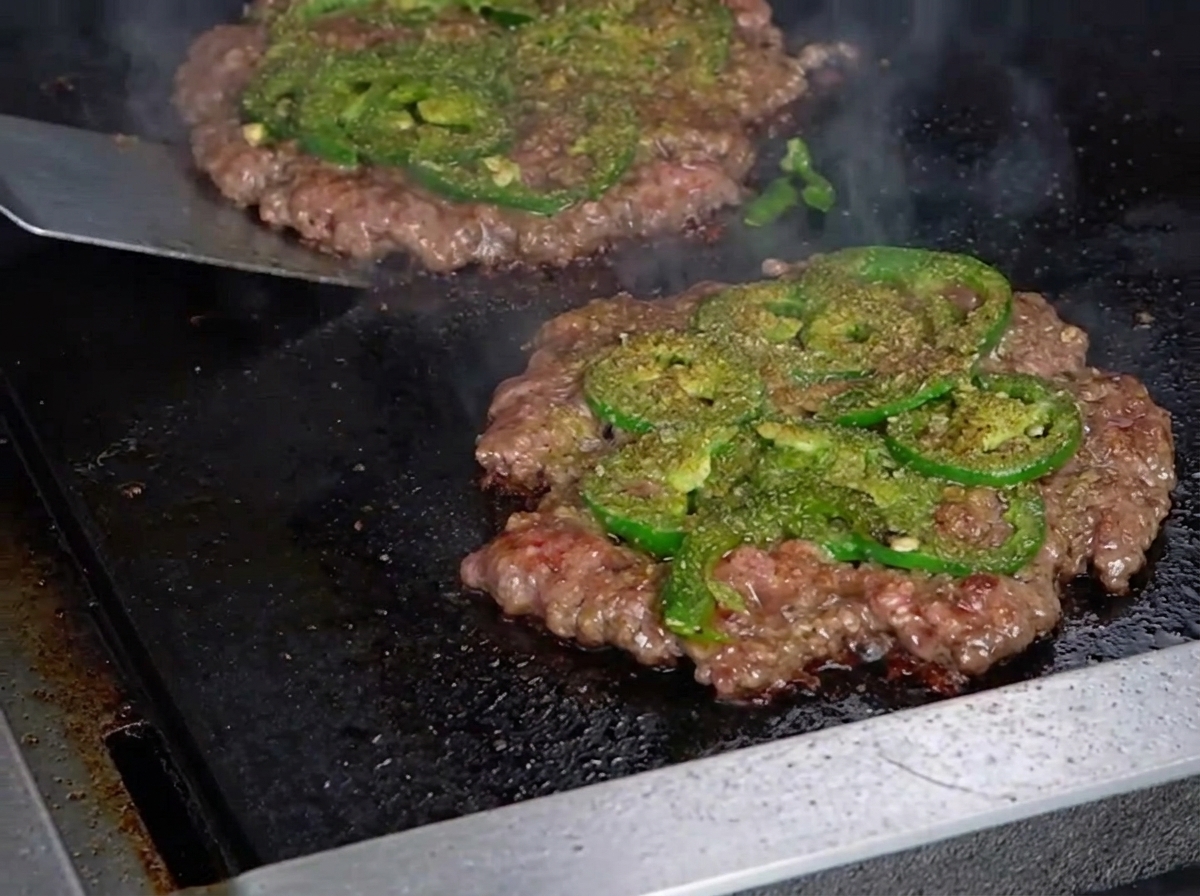 Une spatule en métal retournant un fin steak de smashburger, révélant une belle croûte caramélisée et croustillante sur le dessous.