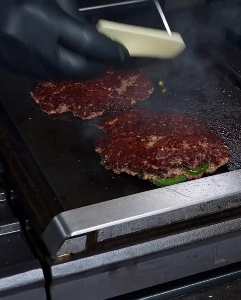 Une main gantée déposant une tranche de fromage Havarti blanc sur un steak de smashburger magnifiquement saisi.