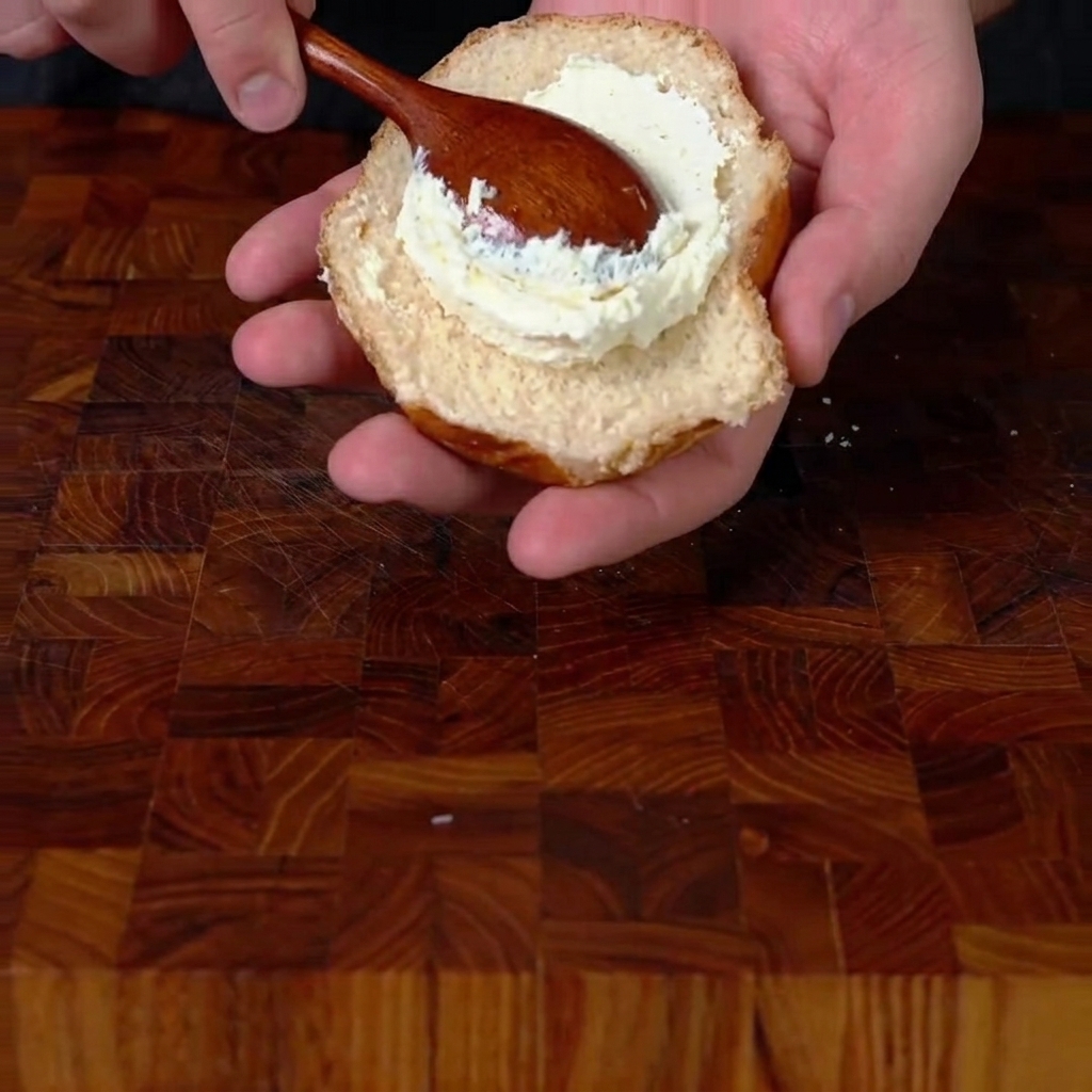 Utilisation dune cuillère en bois pour étaler une épaisse couche de fromage à la crème fouetté sur la moitié inférieure dun pain à burger grillé.