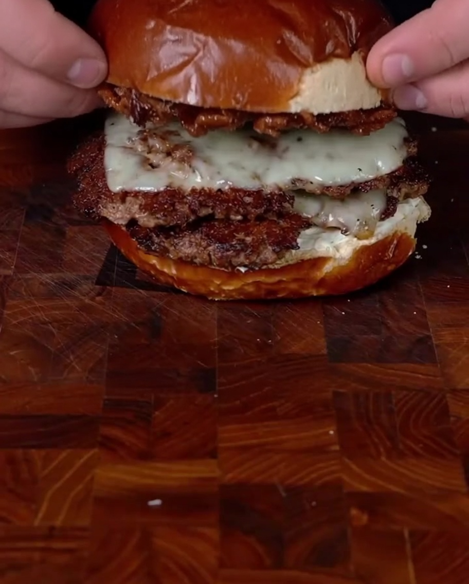 Des mains plaçant le pain du haut chargé de confiture de bacon sur le double smashburger empilé pour terminer lassemblage.