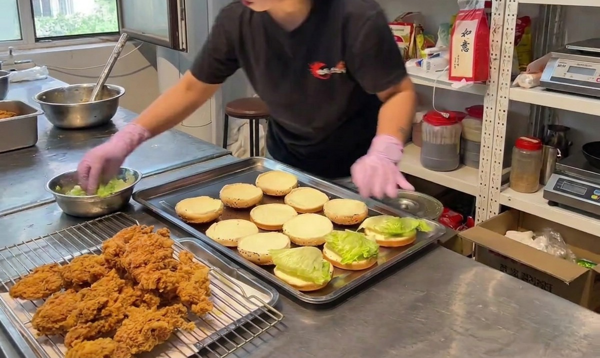 Une personne plaçant un morceau de poulet frit croustillant sur un pain burger inférieur garni de laitue verte.