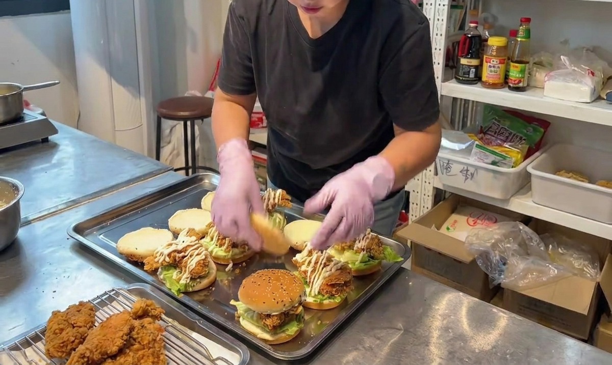 Une personne plaçant la moitié supérieure dun pain aux graines de sésame sur un burger au poulet frit entièrement assemblé.