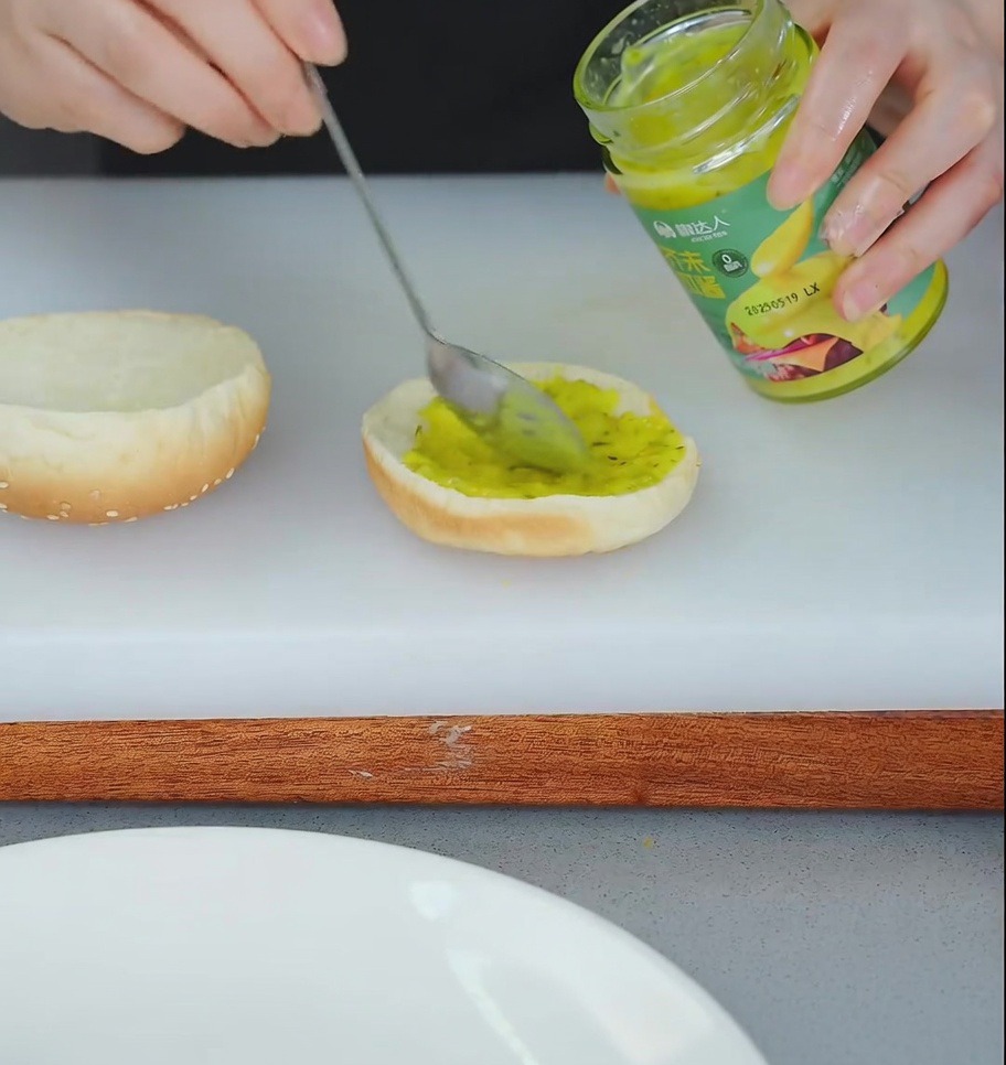 Une personne utilisant une cuillère pour étaler de la relish jaune sur la base dun pain à burger grillé.