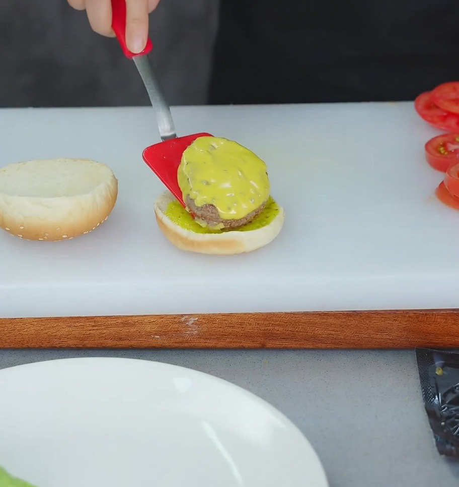 Une spatule rouge déposant un steak au fromage sur la base dun pain à burger nappée de relish.