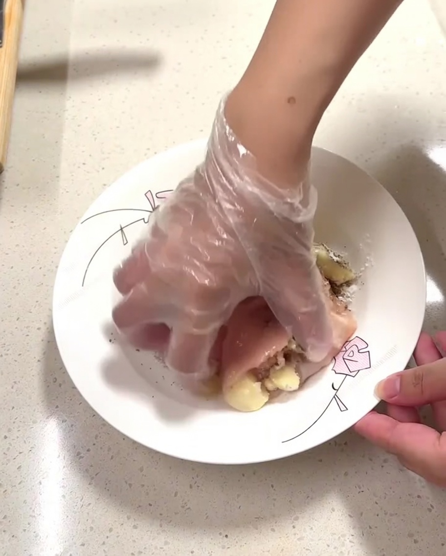 Une main gantée massant les assaisonnements sur les morceaux de poulet cru dans une assiette.