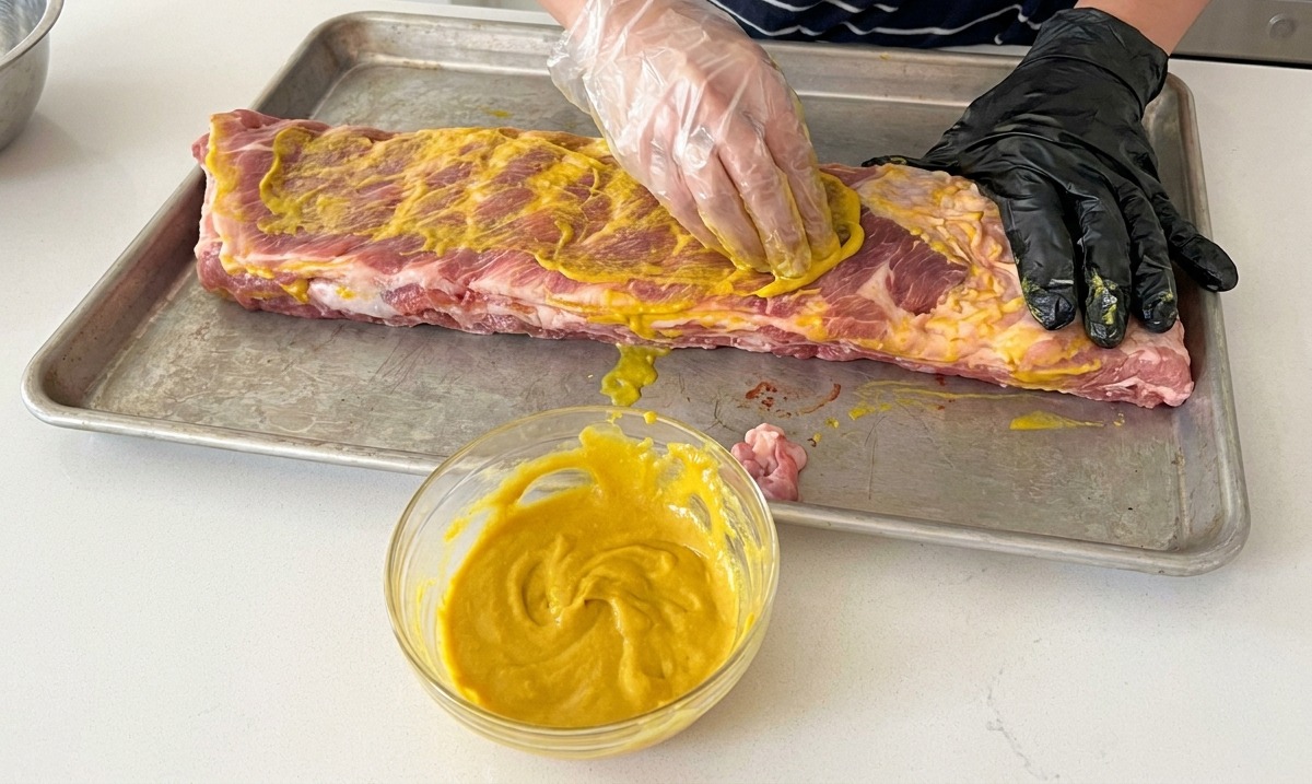 Une personne portant des gants appliquant un liant à la moutarde jaune sur des travers de porc crus.