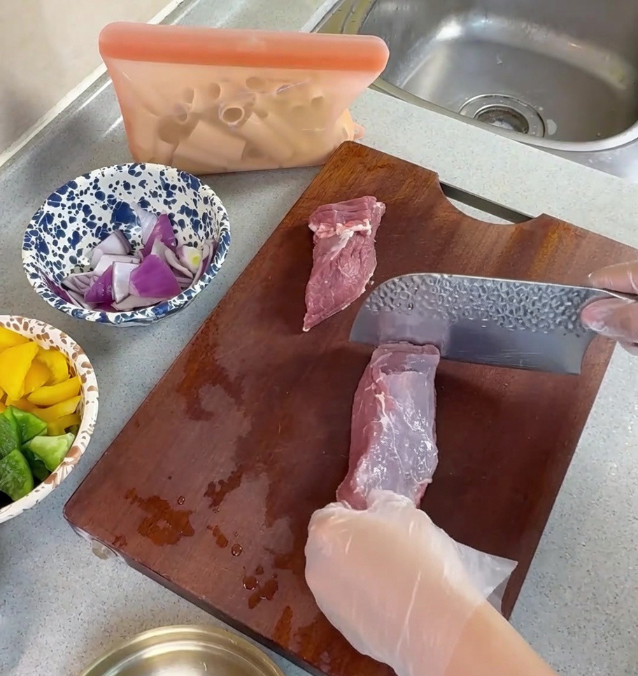 Mains gantées utilisant un couperet pour trancher du porc cru en fines lanières sur une planche à découper en bois.