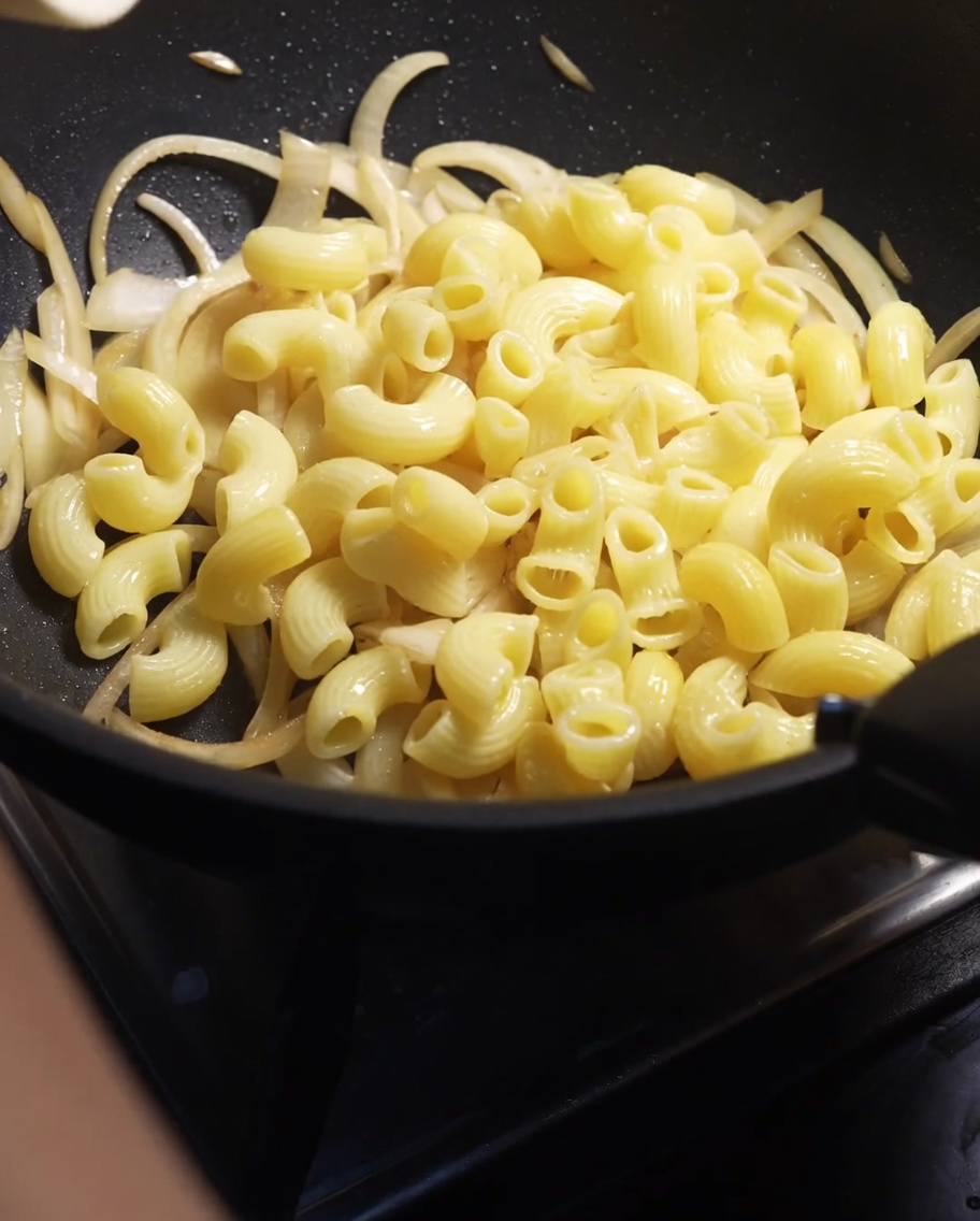 Macaronis cuits versés dans une poêle avec des oignons émincés ramollis.