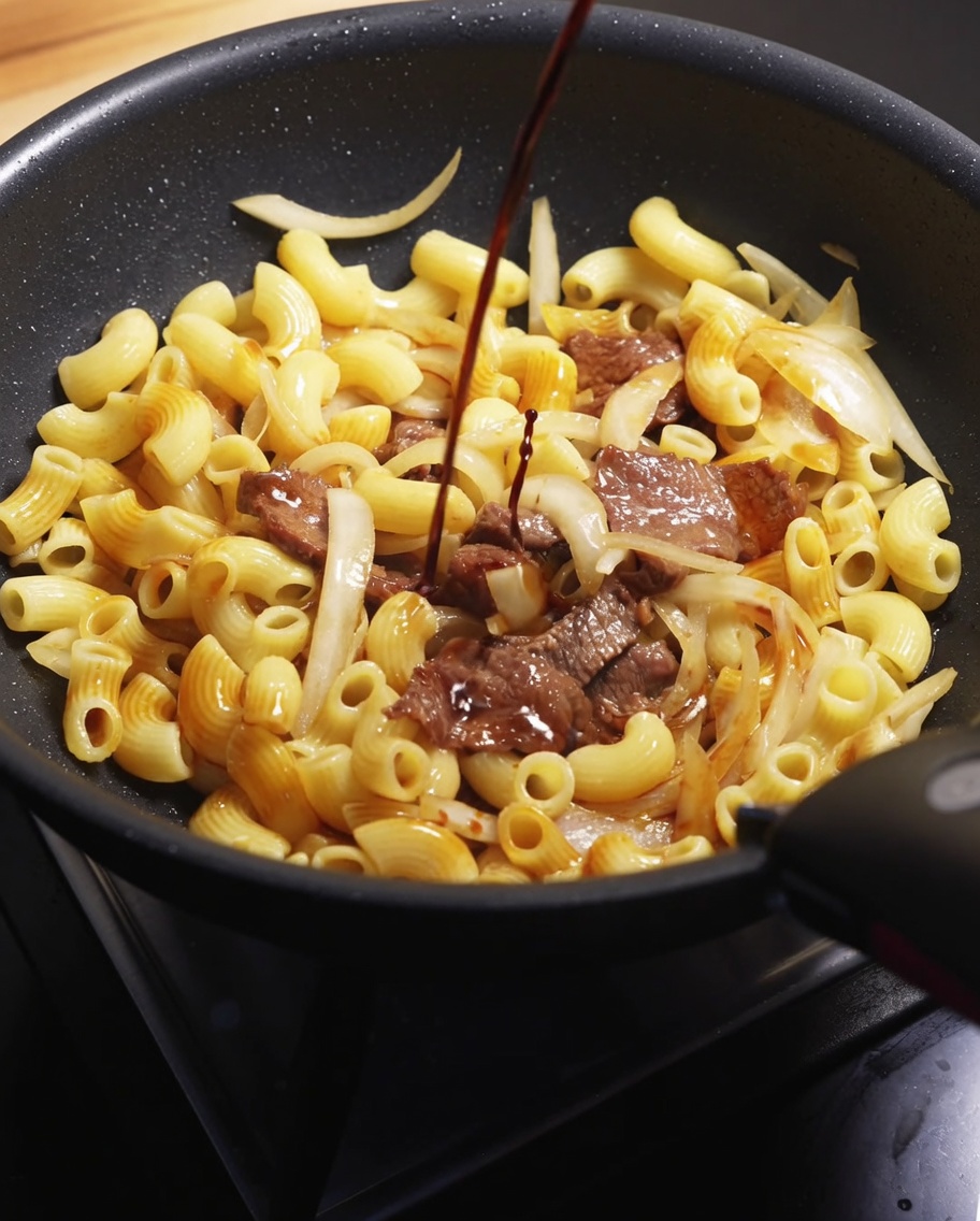 Un filet de sauce soja foncée versé sur le mélange de bœuf et de macaronis dans une poêle chaude.