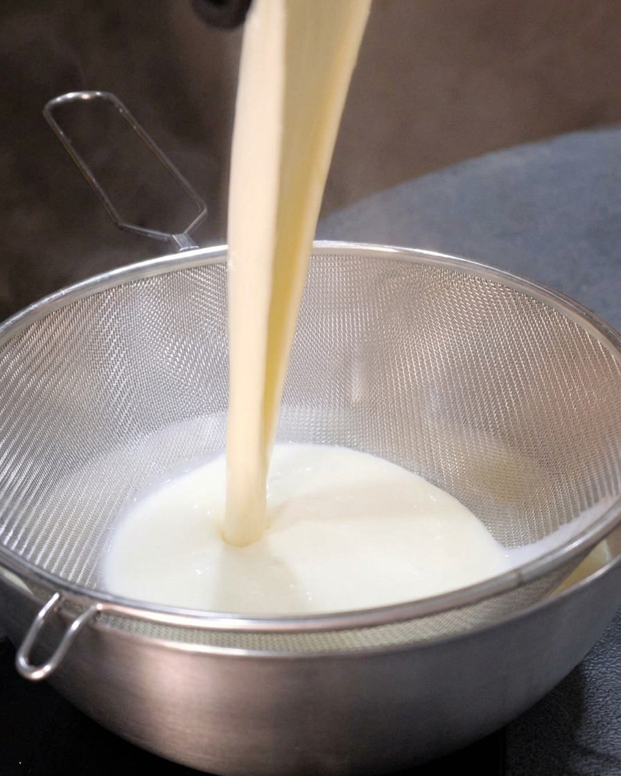 Base de soupe blanche versée dans un tamis en métal, donnant un liquide lisse dans le bol en dessous.