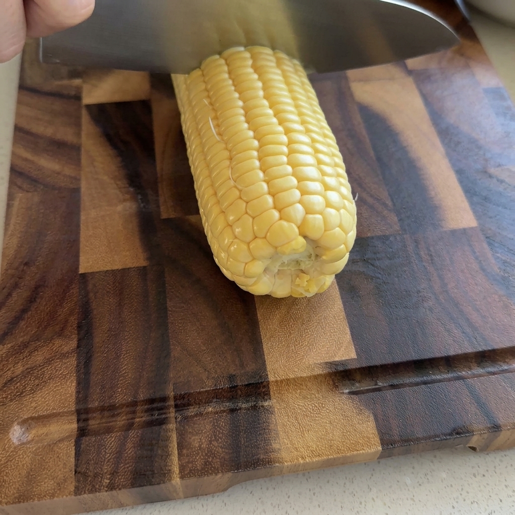 Une main utilisant un grand couteau de chef pour trancher les grains de maïs jaune crus sur une planche à découper en bois.