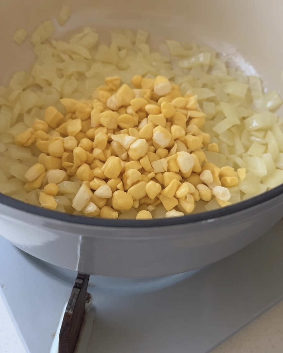 Grains de maïs jaune frais versés dans une casserole contenant des oignons translucides.