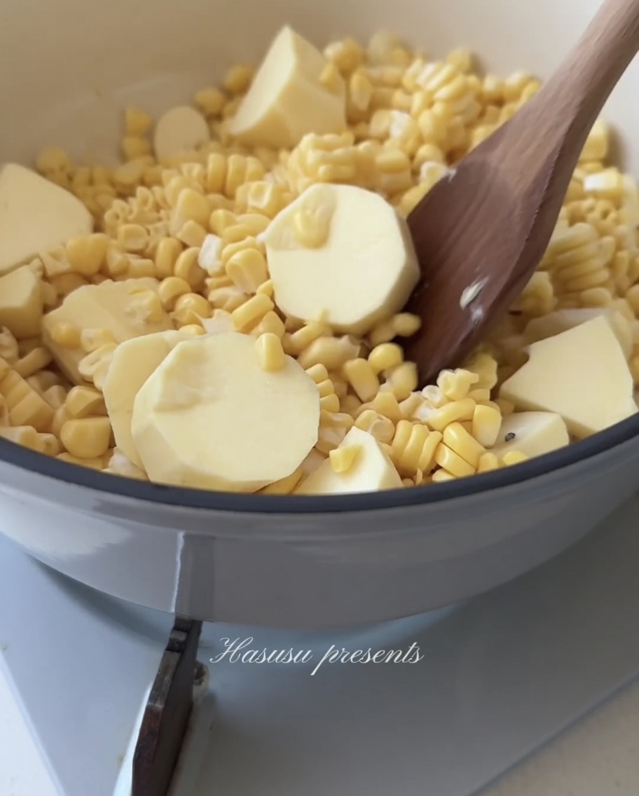 Une cuillère en bois remuant un mélange de maïs et de morceaux de patate douce blanche dans une casserole.