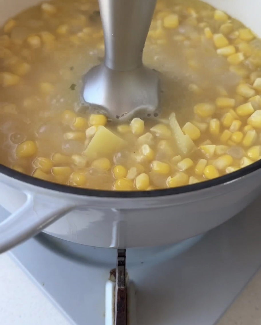 Un mixeur plongeant immergé dans une marmite, mixant les grains de maïs et la patate douce en une purée lisse.