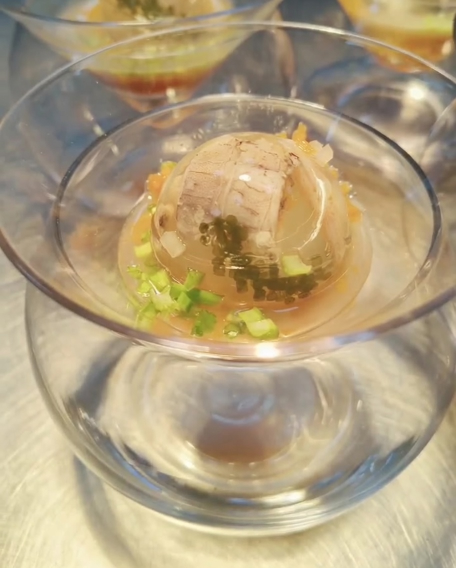 Une sphère de gelée de fruits de mer parfaitement prise dans un bol en verre, arrosée dune riche sauce ambrée, prête à être garnie et servie.