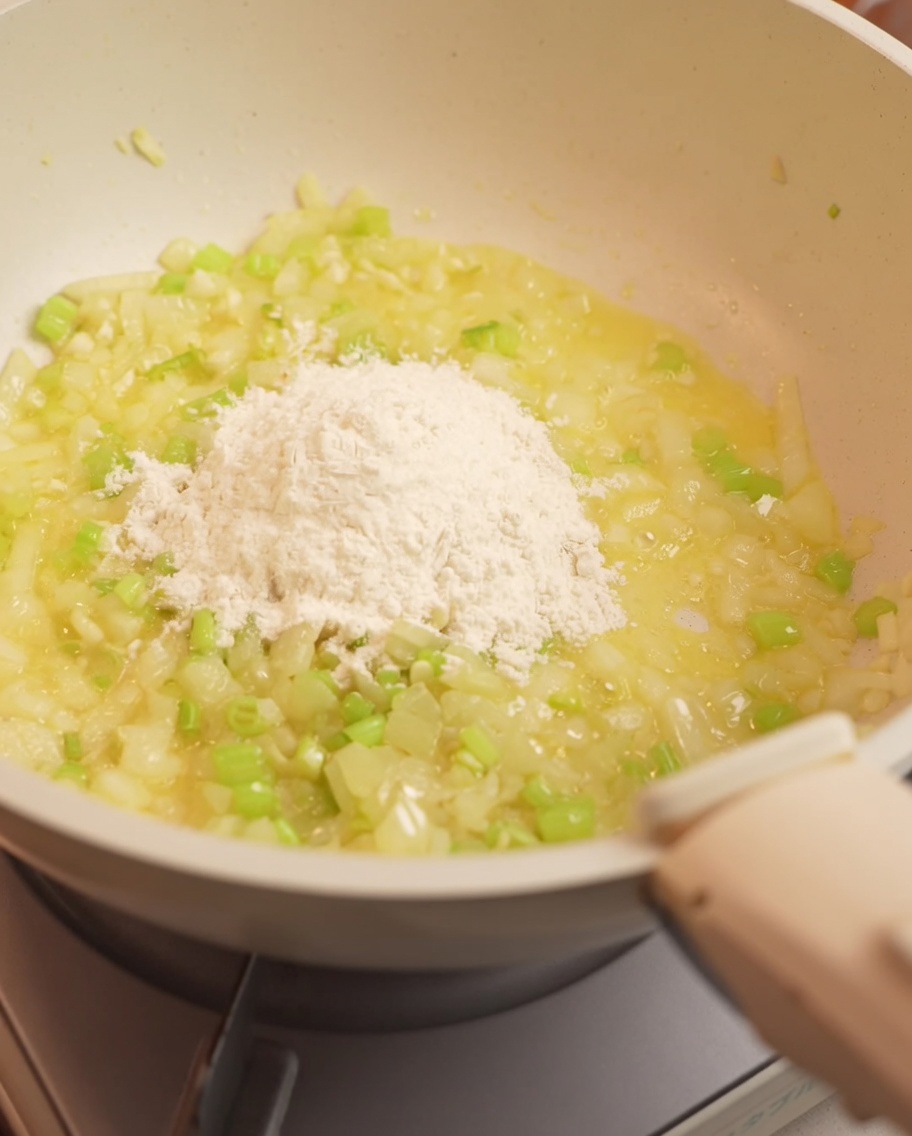 Farine saupoudrée et incorporée au mélange doignons et de céleri dans une poêle.
