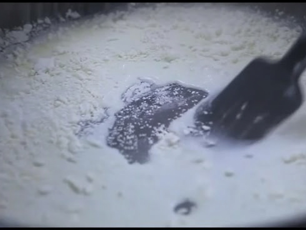 Une spatule en silicone noire remuant un mélange épais et blanc de lait et de farine dans une poêle.