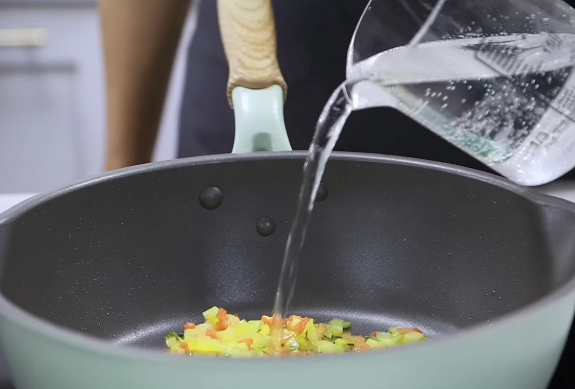 De leau versée dun verre doseur dans une poêle contenant des légumes poêlés, en préparation de lincorporation du mélange de lait épaissi.