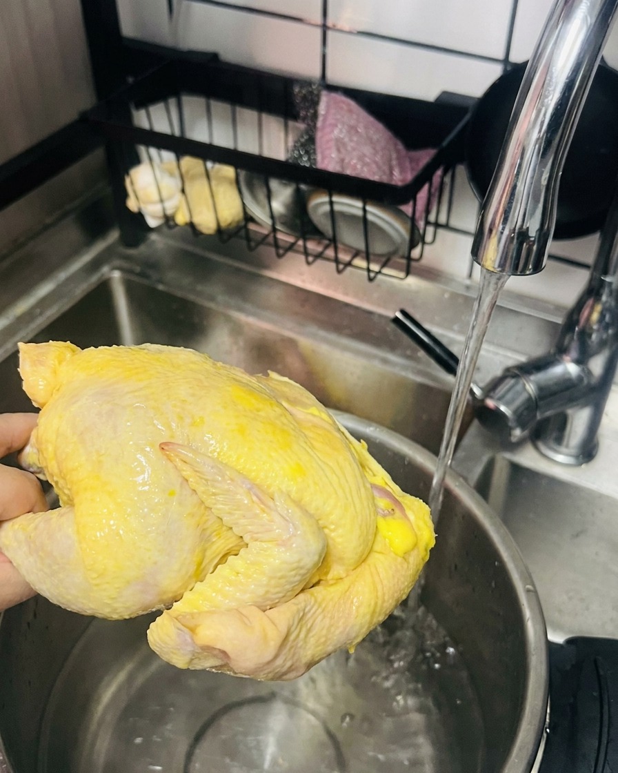 Une main tenant une poule jaune crue entière sous un robinet deau, prête à être placée dans une marmite.