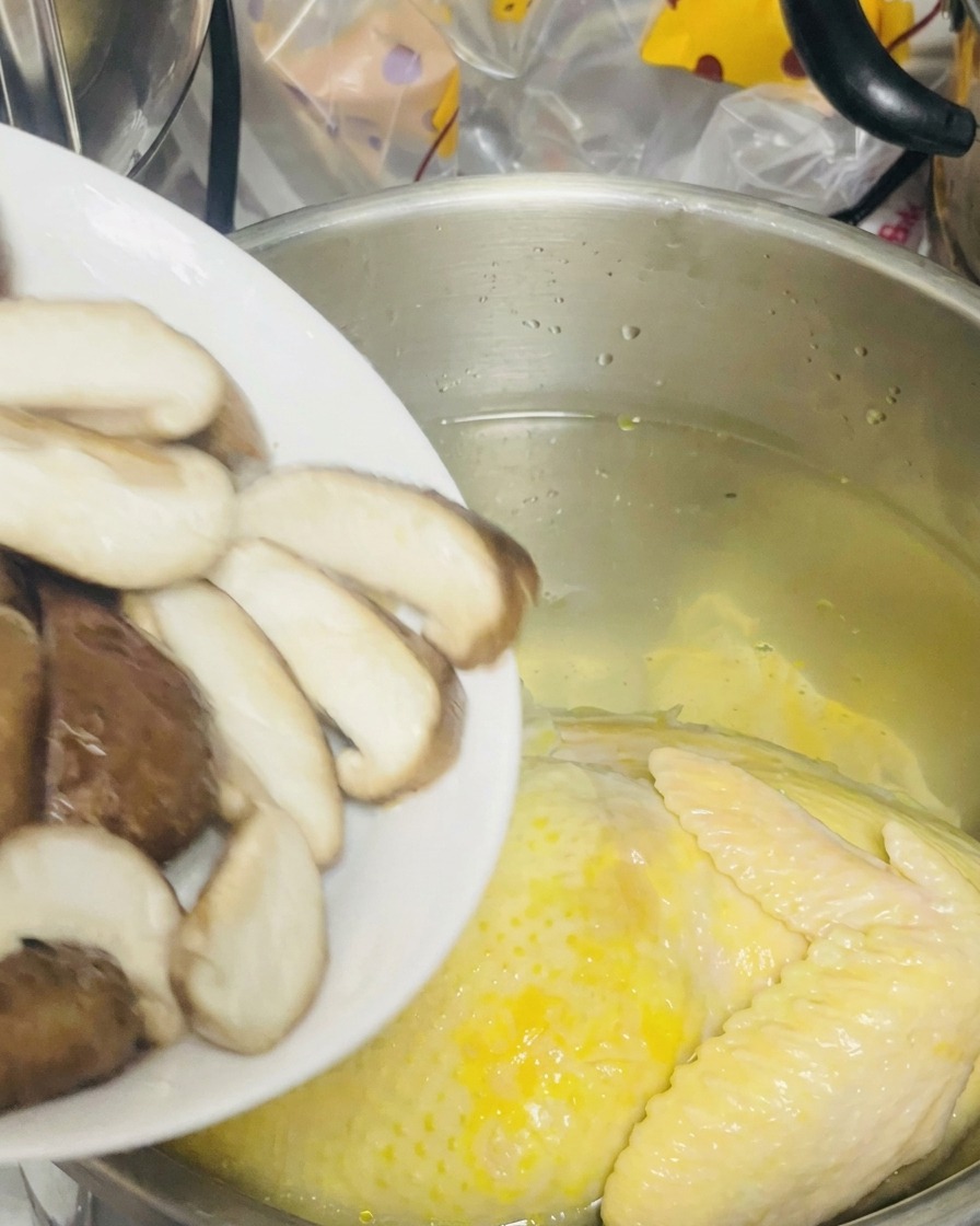 Des shiitakés émincés versés depuis une assiette blanche dans une marmite contenant une poule dans leau.