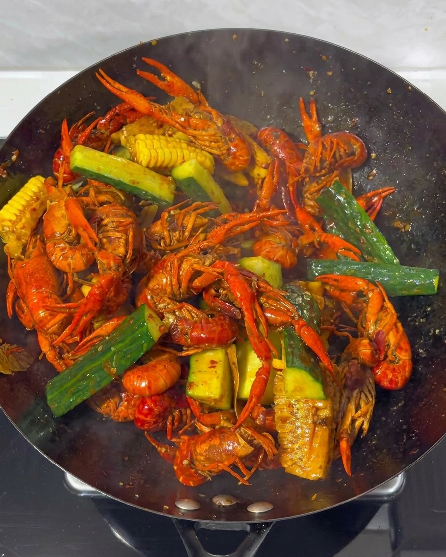Gros plan sur les écrevisses, le maïs et le concombre mélangés dans un grand wok avec une sauce rouge foncé.