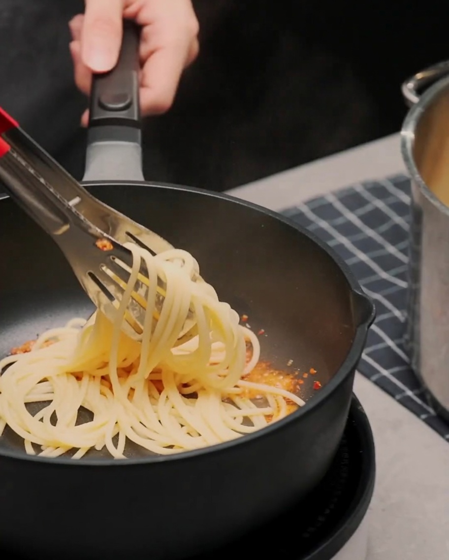 Spaghetti cuits mélangés à laide de pinces dans une poêle avec lail et lhuile pimentée.