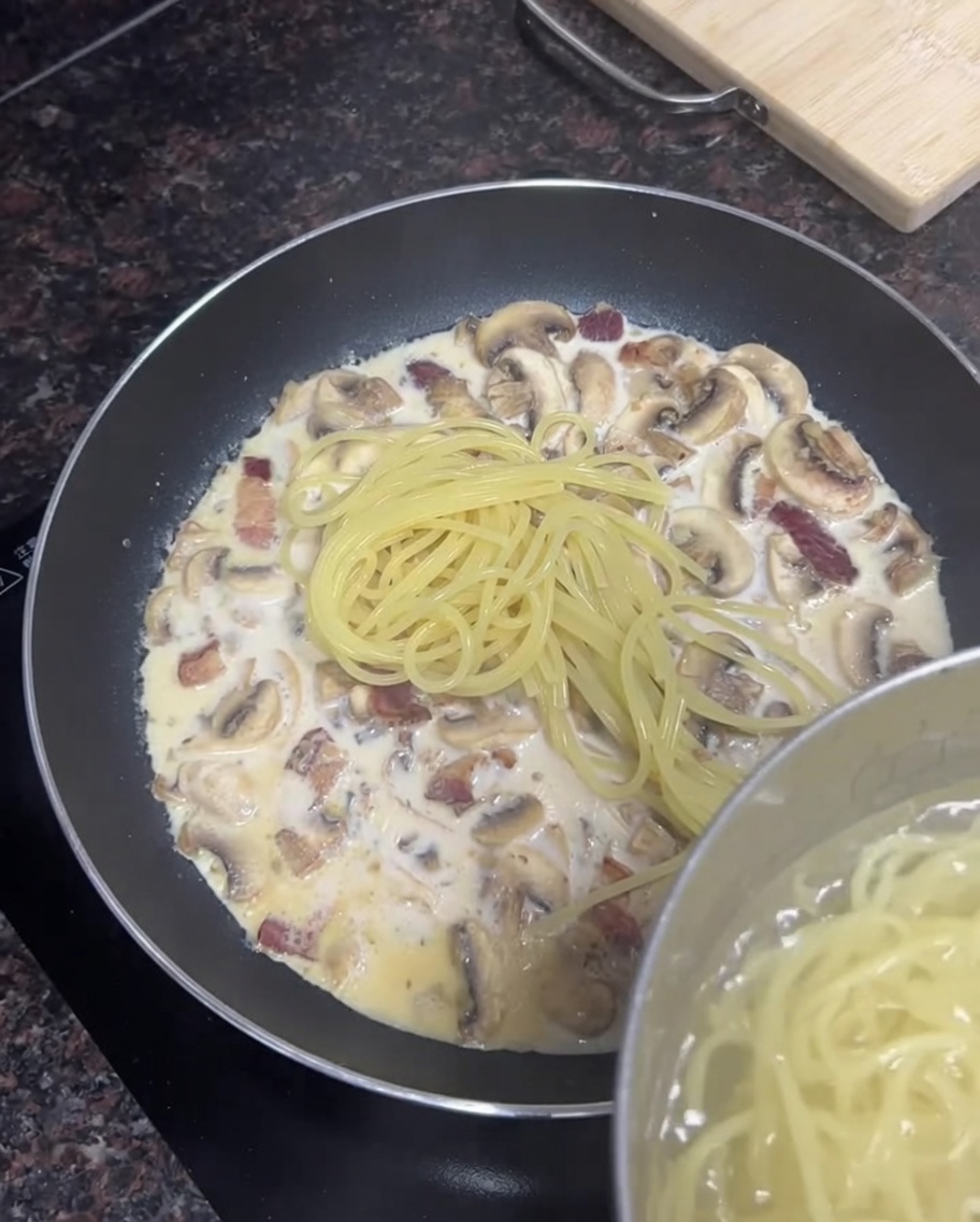 Spaghettis cuits ajoutés depuis une casserole dans une poêle remplie de sauce crémeuse au bacon et aux champignons.