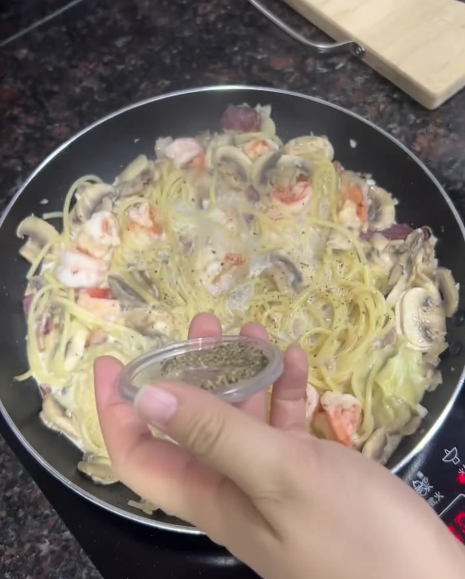 Une main saupoudrant du poivre noir fraîchement moulu dun petit récipient sur une poêle de pâtes crémeuses aux crevettes et champignons.