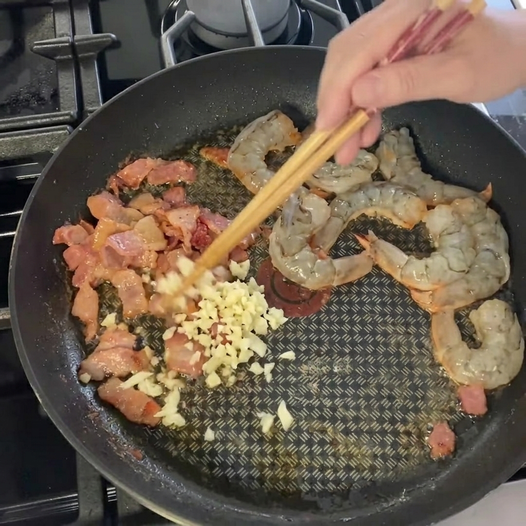 Ail haché ajouté aux crevettes et au lard dans la poêle.