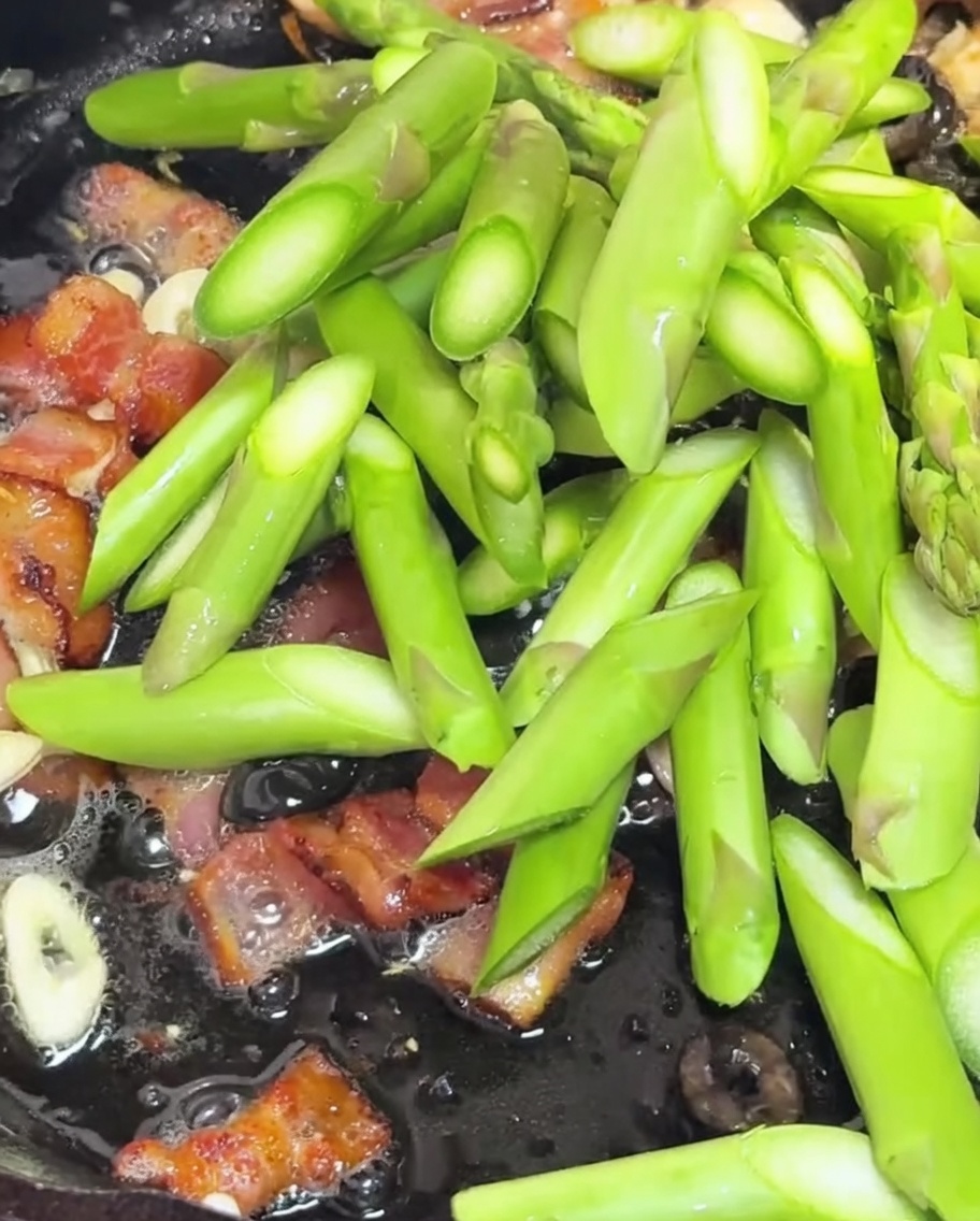 Des tronçons dasperges et des tomates cerises coupées en deux sautant dans une poêle avec le bacon et lail.