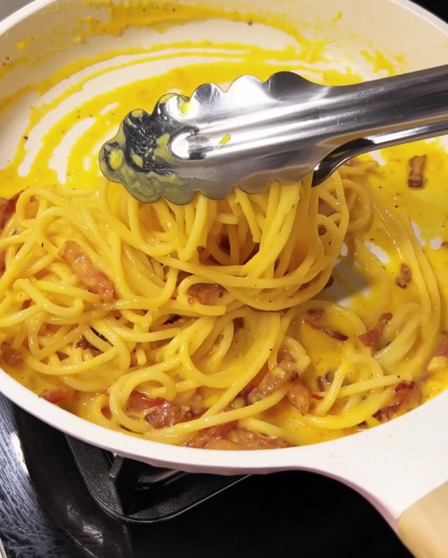 Pinces mélangeant les spaghetti, la sauce et le guanciale dans la poêle.