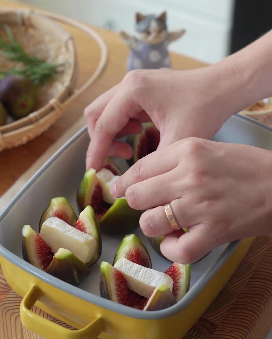 Moitiés de figues dans un plat de cuisson jaune, en cours de garnissage avec des morceaux de Brie.